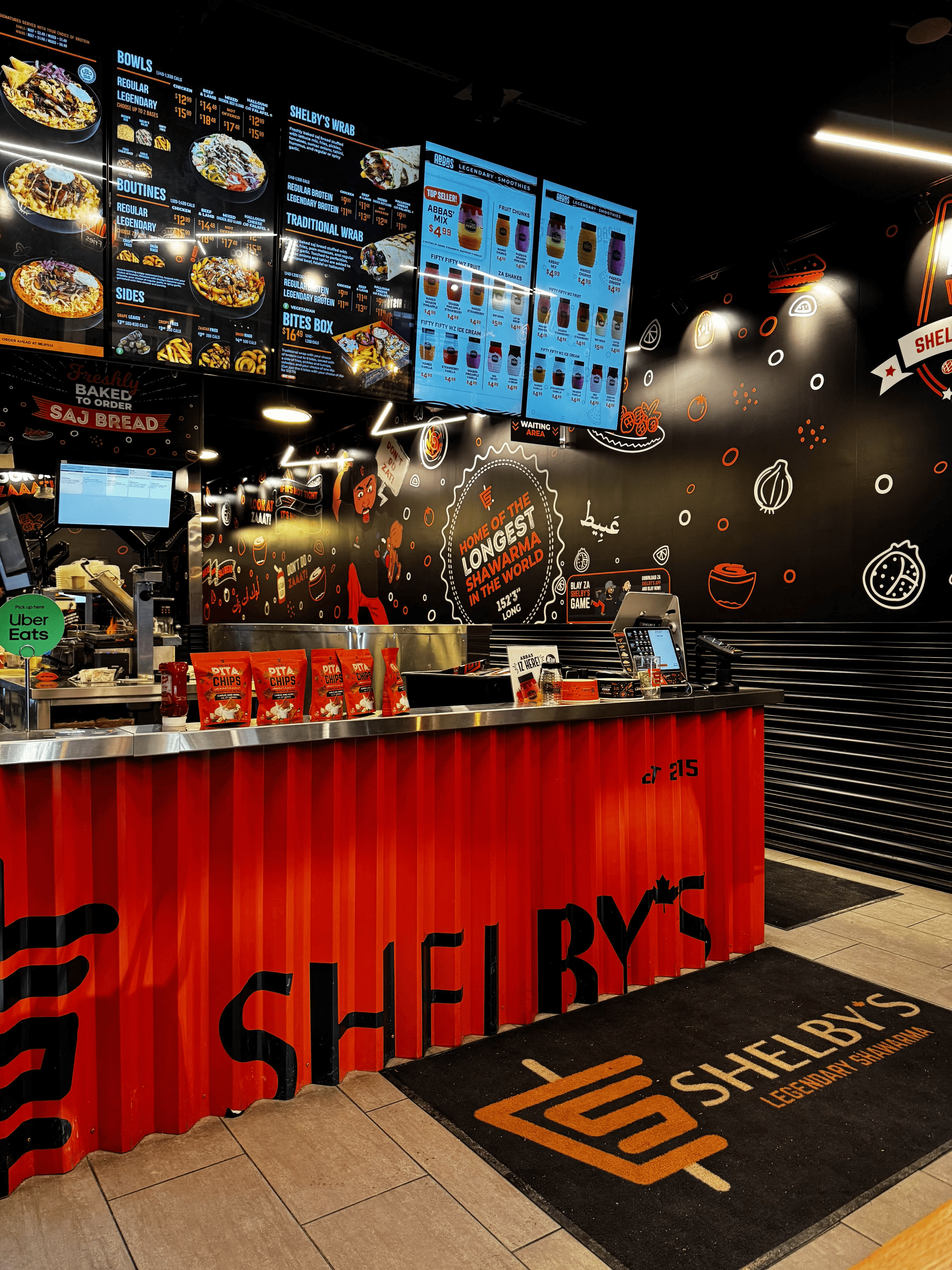 Inside a Shelby’s Legendary Shawarma restaurant: digital menu boards above a red corrugated counter, an illustrated black wall and a Shelby’s floor mat.
