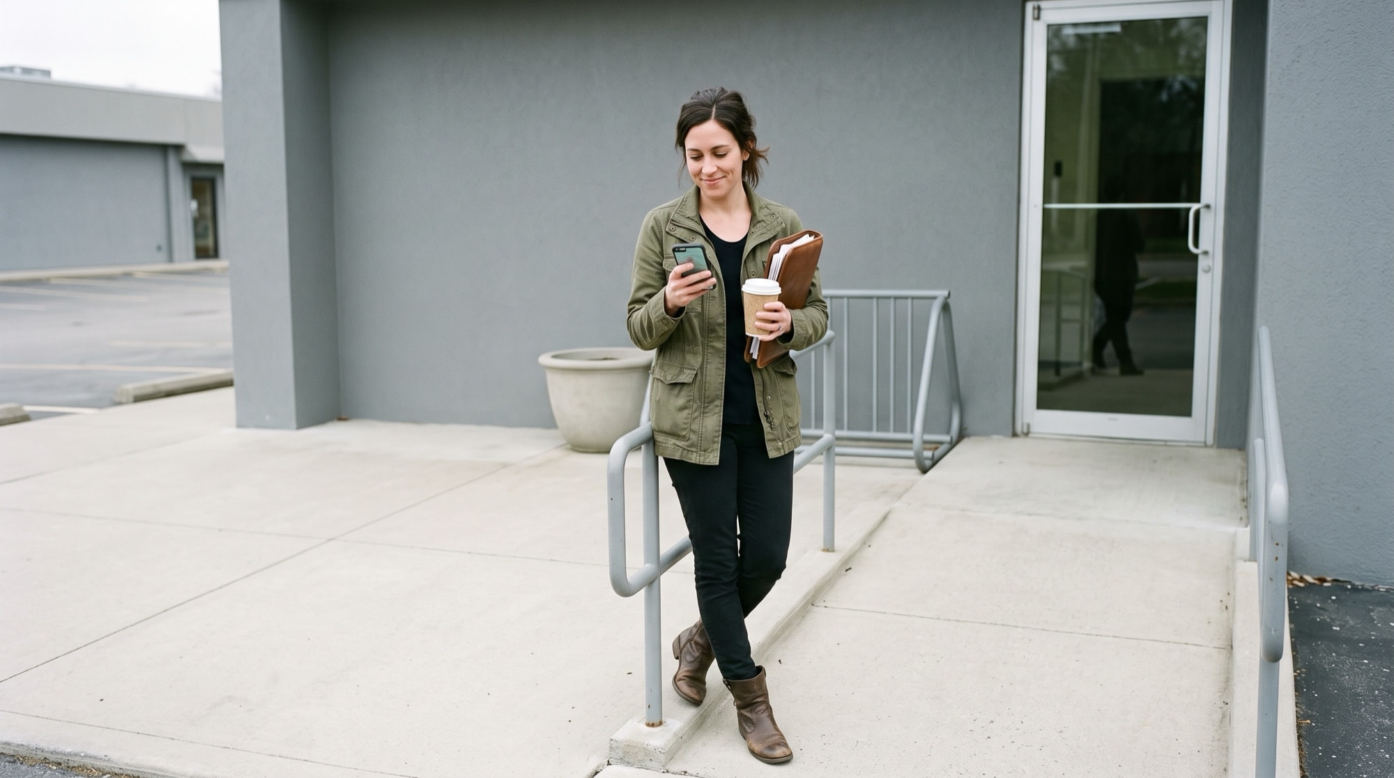 A woman pauses outside an ordinary commercial building with a phone in one hand and a coffee cup in the other, tapping a single approval on the screen.